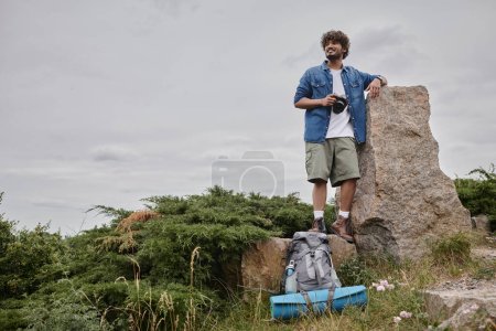 photography and nature concept, happy indian backpacker holding digital camera and standing on rock