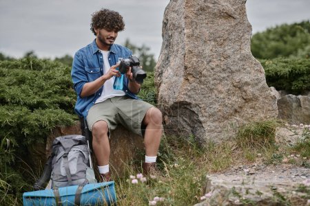 Téléchargez les photos : Voyage et concept de la nature, homme indien tenant bouteille et en utilisant un appareil photo numérique tout en étant assis sur le roc - en image libre de droit