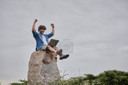 digitales Nomadenkonzept, aufgeregter indischer Freelancer, der den Sieg feiert, während er Laptop benutzt, auf Felsen sitzend