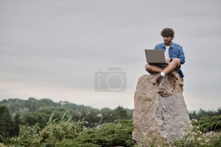 digitaler Nomade, lockiger indischer Mann mit Laptop, auf Felsen sitzend, Fernarbeitskonzept, Landschaftsansicht