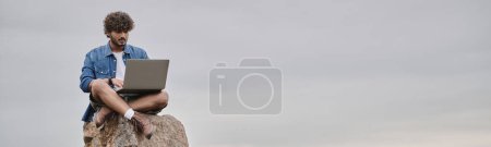 Téléchargez les photos : Nomade numérique, homme indien concentré utilisant un ordinateur portable, assis sur le rocher, concept de travail à distance, bannière - en image libre de droit