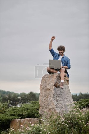 digitales Nomadenkonzept, aufgeregter Indianer, der den Sieg feiert, während er Laptop benutzt, sitzt auf einem Felsen
