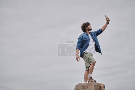 Foto de Concepto de nomadismo, hombre indio rizado de pie en el rock y la señal de búsqueda mientras sostiene el teléfono inteligente - Imagen libre de derechos