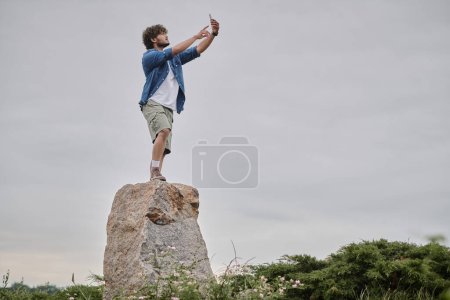 Foto de Concepto de nomadismo, el hombre indio rizado de pie en el rock y la señal de búsqueda mientras sostiene el teléfono móvil - Imagen libre de derechos