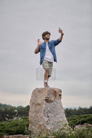 nomadism concept, curly indian man standing on rock and having video chat on smartphone, wave hand