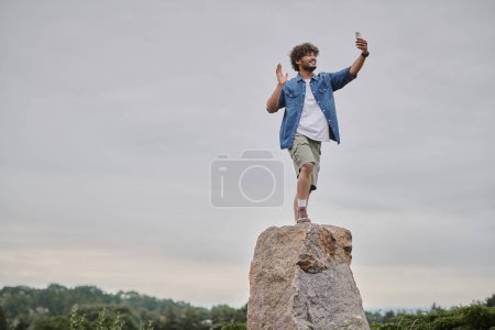 nomadism concept, happy indian man standing on rock and having video chat on smartphone, wave hand