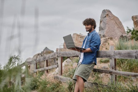 concepto nómada digital, hombre indio alegre de pie cerca de valla de madera y el uso de ordenador portátil, la naturaleza