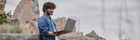 digital nomad concept, cheerful indian man standing near wooden fence and using laptop, banner
