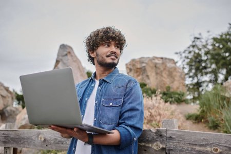 digital nomad concept, positive indian man standing near wooden fence and using laptop, countryside