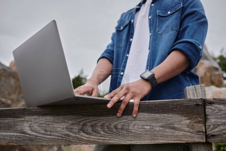 cropped view of freelancer standing near wooden fence and using laptop, digital nomad concept