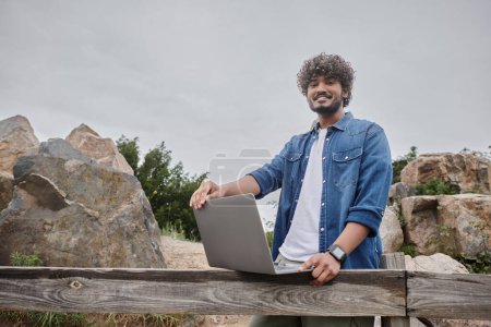positiver indischer Mann, der in der Nähe eines Holzzaunes in der Landschaft steht und Laptop, digitales Nomadentum benutzt