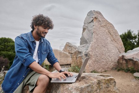Photo for Happy indian man typing on laptop and working remotely in natural place, digital nomadism - Royalty Free Image