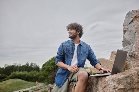 cheerful indian freelancer working remotely and using laptop in natural place, digital nomadism