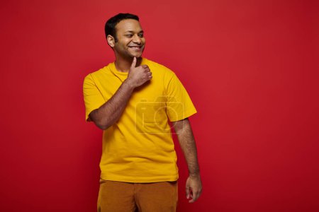 glücklicher indischer Mann in heller Freizeitkleidung stehend und lächelnd auf rotem Hintergrund im Studio
