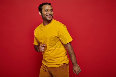 Téléchargez les photos : Homme indien joyeux en vêtements décontractés lumineux regardant loin et souriant sur fond rouge, optimiste - en image libre de droit