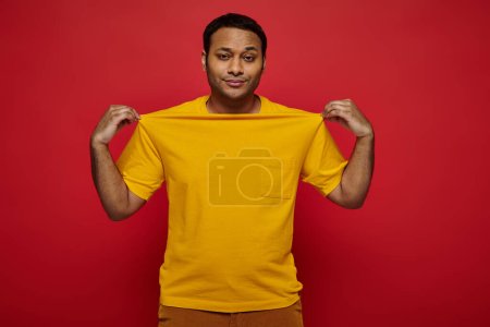 modischer indischer Mann, der gelbes T-Shirt berührt und vor rotem Hintergrund in die Kamera schaut, persönlicher Stil