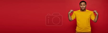 fashionable indian man touching yellow t-shirt and looking at camera on red backdrop, banner