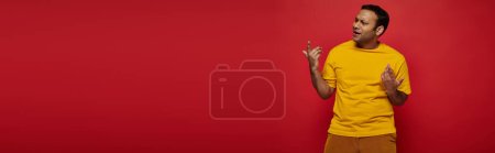 Téléchargez les photos : Homme indien émotionnel en vêtements décontractés lumineux gestuelle tout en expliquant quelque chose sur rouge, bannière - en image libre de droit
