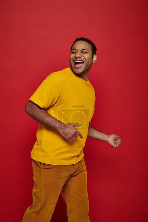 hombre indio excitado en ropa casual brillante sonriendo y mirando hacia otro lado en el fondo rojo en el estudio