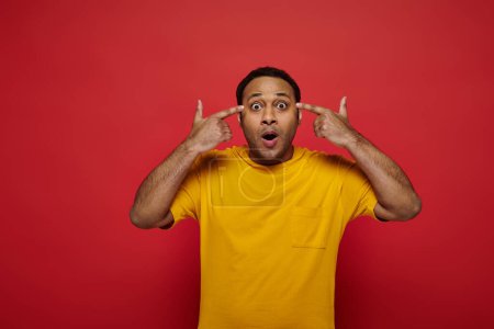Photo for Mind blowing, shocked indian man with fingers near head looking at camera on red backdrop in studio - Royalty Free Image