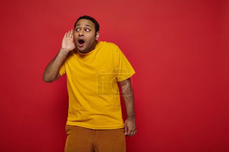 Photo for Curious indian man in bright clothes looking away and listening on red backdrop, hand near ear - Royalty Free Image