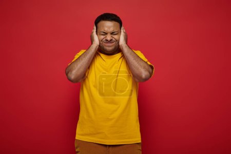 tensed indian man in bright clothes standing with closed eyes and covering ears on red background