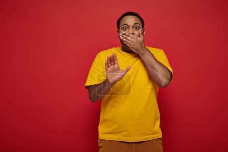 shocked indian man in bright clothes covering mouth and showing stop sign on red background