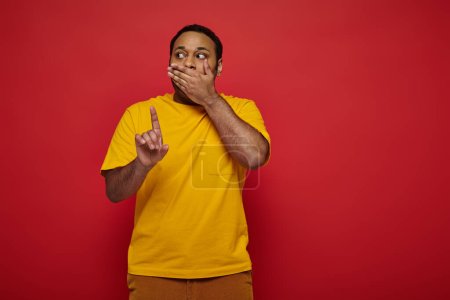 schockierter indischer Mann in heller Kleidung, der den Mund bedeckt und den Finger auf rotem Hintergrund zeigt, warnt