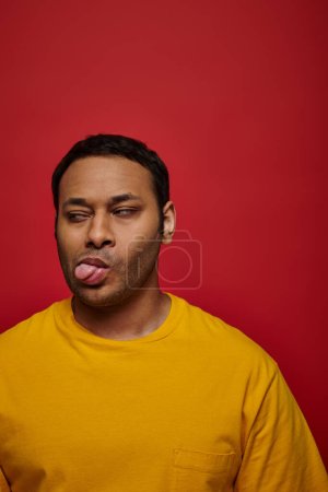 Gesichtsausdruck, unzufriedener indischer Mann in gelber Kleidung, der die Zunge auf rotem Hintergrund herausstreckt