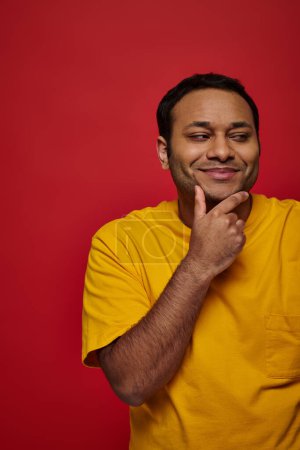 zufriedener indischer Mann im gelben T-Shirt, der sein Kinn berührt und auf rotem Hintergrund wegschaut, nachdenklich