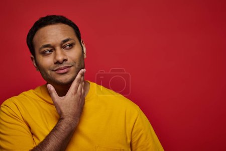 hombre indio soñador en camiseta amarilla tocando la barbilla y mirando hacia otro lado sobre el fondo rojo, reflexivo