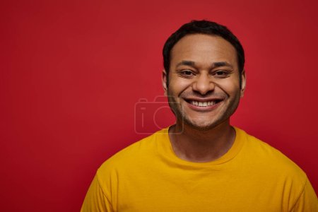 emoción positiva, hombre indio feliz en camiseta amarilla sonriendo o sonriendo sobre fondo rojo en el estudio