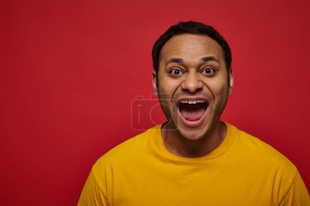 Photo pour Émotion positive, excité homme indien en t-shirt jaune avec bouche ouverte sur fond rouge - image libre de droit