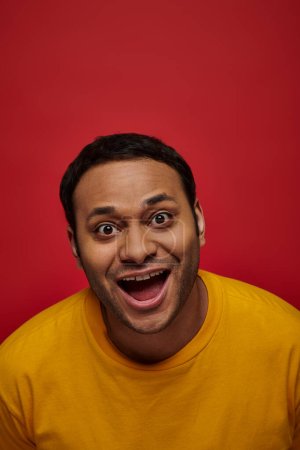 Photo pour Émotion positive, homme indien excité en t-shirt jaune avec bouche ouverte sur fond rouge, vertical - image libre de droit