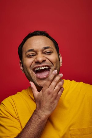 emoción positiva, hombre indio excitado en camiseta amarilla riendo con la boca abierta sobre fondo rojo