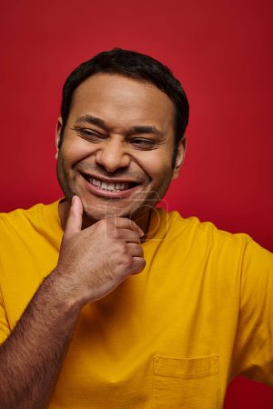 emoción positiva, hombre indio alegre en camiseta amarilla sonriendo y tocando la barbilla sobre fondo rojo