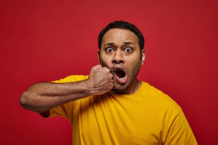 expresión de la cara, impactado hombre indio en camiseta amarilla golpeándose en la cara en el fondo rojo