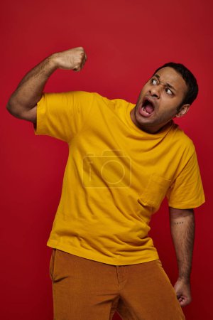 Gesichtsausdruck, verängstigter indischer Mann in gelbem T-Shirt, der sich vor rotem Hintergrund ins Gesicht schlägt