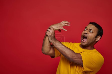 Gesichtsausdruck, verängstigter indischer Mann in gelbem T-Shirt verliert die Kontrolle über seine Hand auf rotem Hintergrund