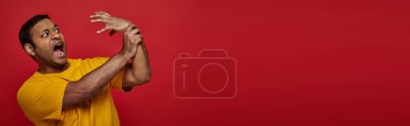 Photo for Face expression, scared indian man in yellow t-shirt losing control of his hand on red background - Royalty Free Image