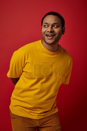 fröhlicher indischer Mann im gelben T-Shirt, der wegschaut und auf rotem Hintergrund lächelt, Gesichtsausdruck
