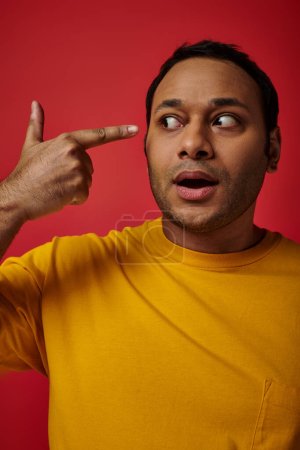 Foto de Impactado hombre indio en camiseta amarilla señalando con el dedo en la cabeza sobre fondo rojo en el estudio - Imagen libre de derechos