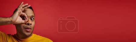 Téléchargez les photos : Homme indien étonné en t-shirt jaune montrant signe correct, main près du visage en arrière-plan en studio, bannière - en image libre de droit