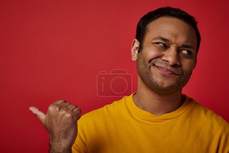 Photo for Skeptical indian man in yellow t-shirt pointing with thumb and looking away on red backdrop - Royalty Free Image