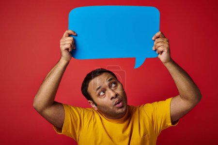 surprised indian man in yellow t-shirt holding blank speech bubble above head on red background