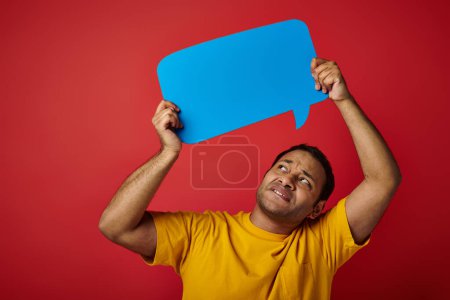 displeased indian man in yellow t-shirt holding blank speech bubble above head on red background