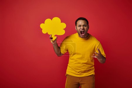 hombre indio enojado en camiseta amarilla sosteniendo burbuja de habla en blanco y gritando sobre fondo rojo