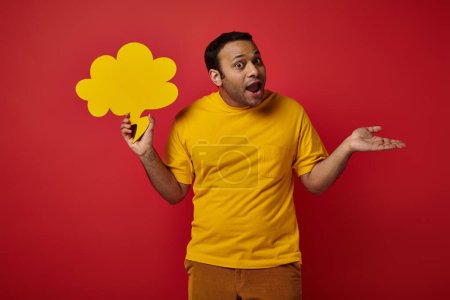 discouraged indian man holding blank speech bubble and showing shrug gesture on red background
