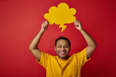 homme indien joyeux en t-shirt jaune tenant bulle de discours vierge au-dessus de la tête sur fond rouge