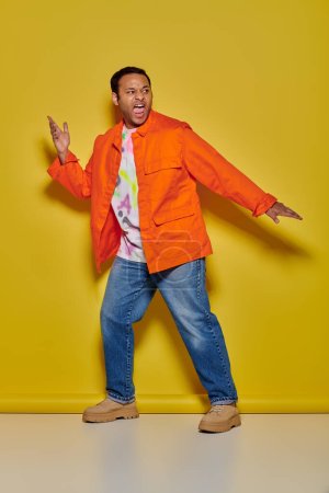 Foto de Hombre indio emocional en chaqueta naranja y jeans haciendo gestos y gritando sobre fondo amarillo - Imagen libre de derechos